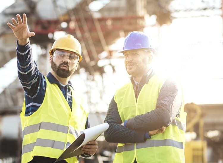 dois trabalhadores de colete amarelo e capacete amarelo e azul discutem as normas regulamentadoras na construção civil